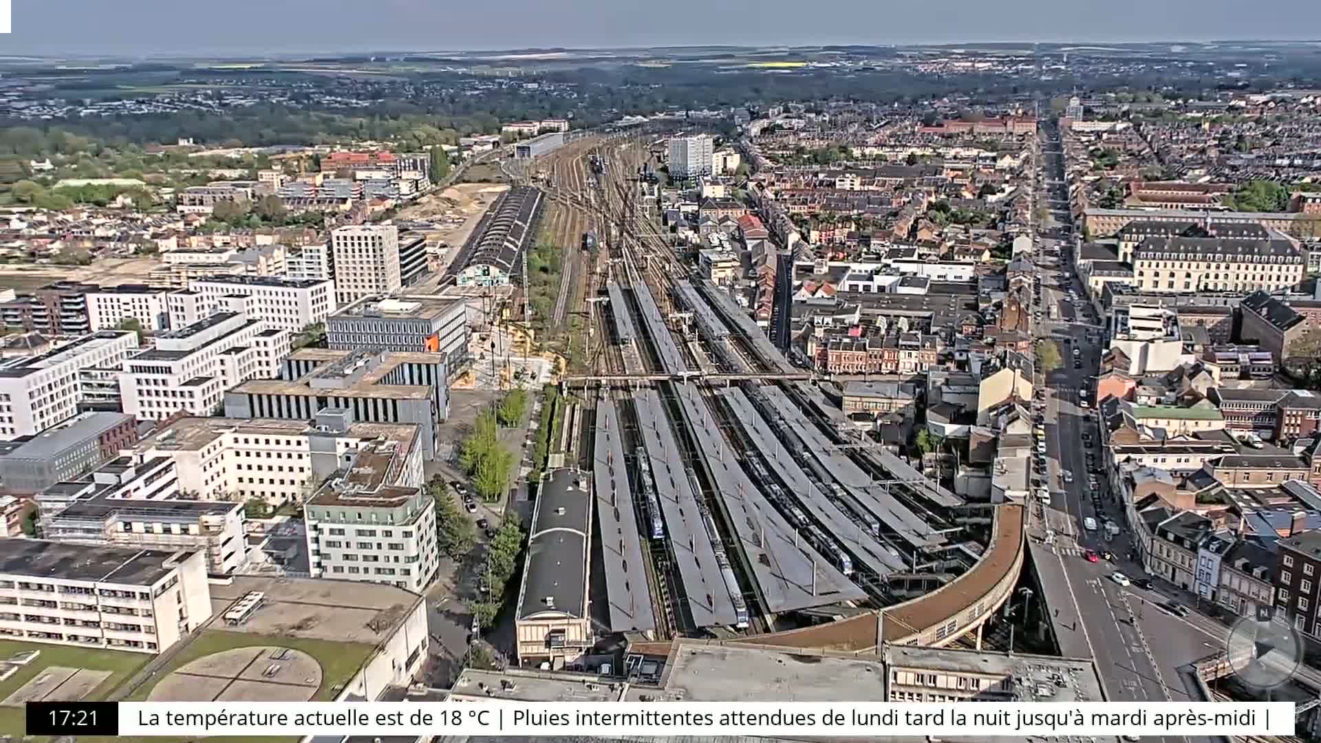 An aerial view of a city featuring a large train station with many tracks and platforms under a mostly clear sky.