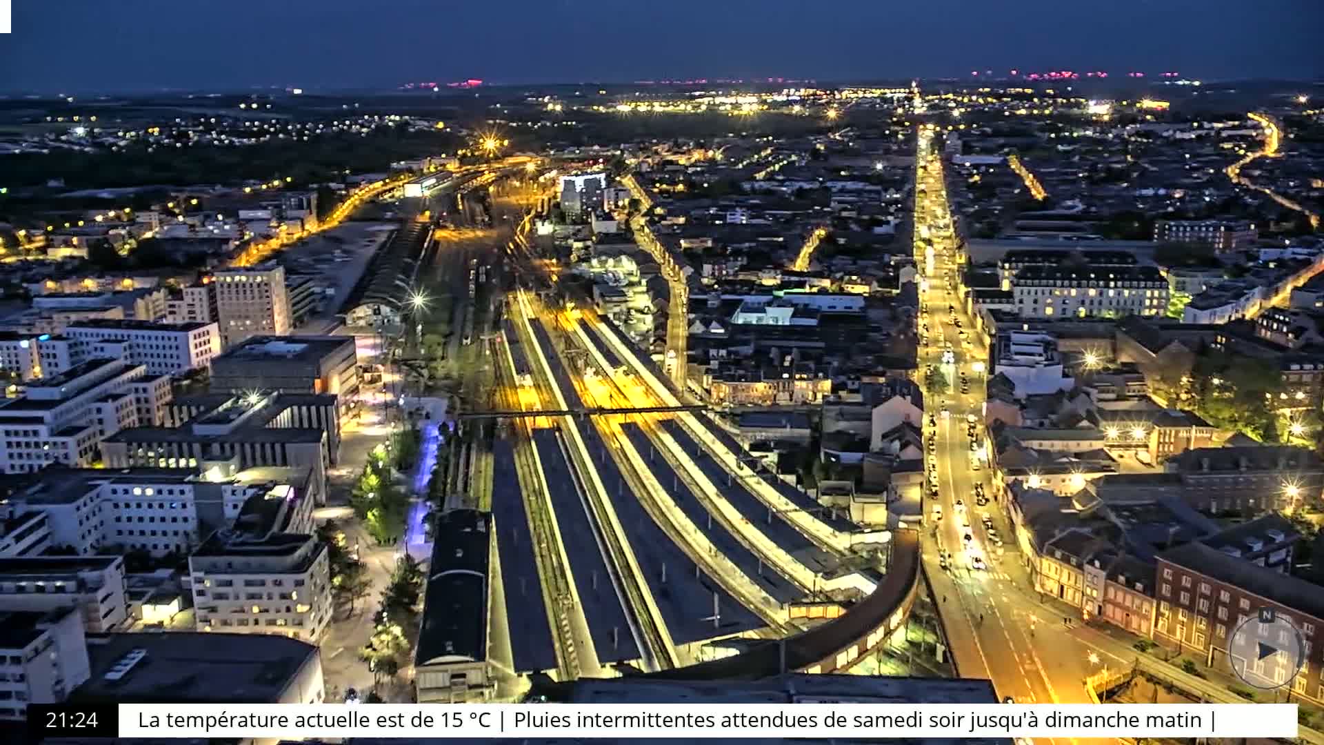 A nighttime aerial view of a city illuminated by streetlights, featuring a large train station and surrounding buildings under a dark sky.