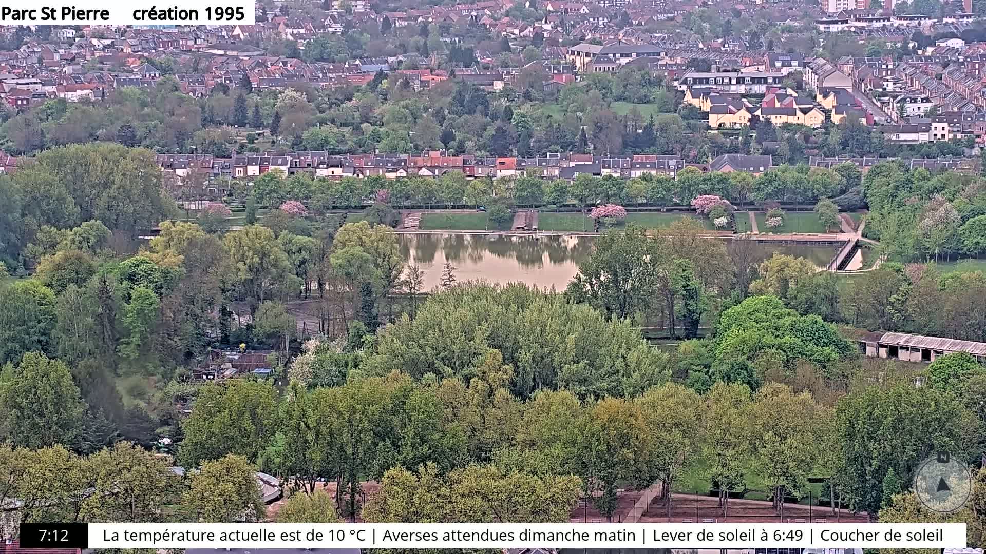 An aerial view shows a park with a calm, reflective pond surrounded by lush green trees and a residential area beyond.