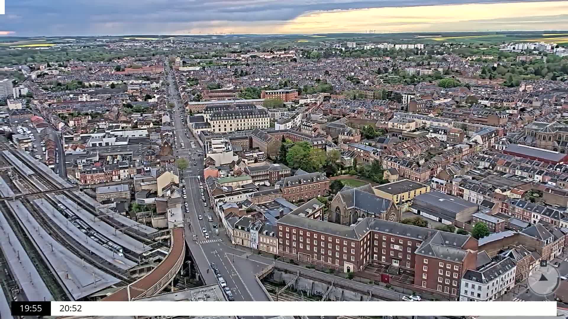 An aerial view shows a densely populated city under partly cloudy skies, with a train station and multiple roads visible.