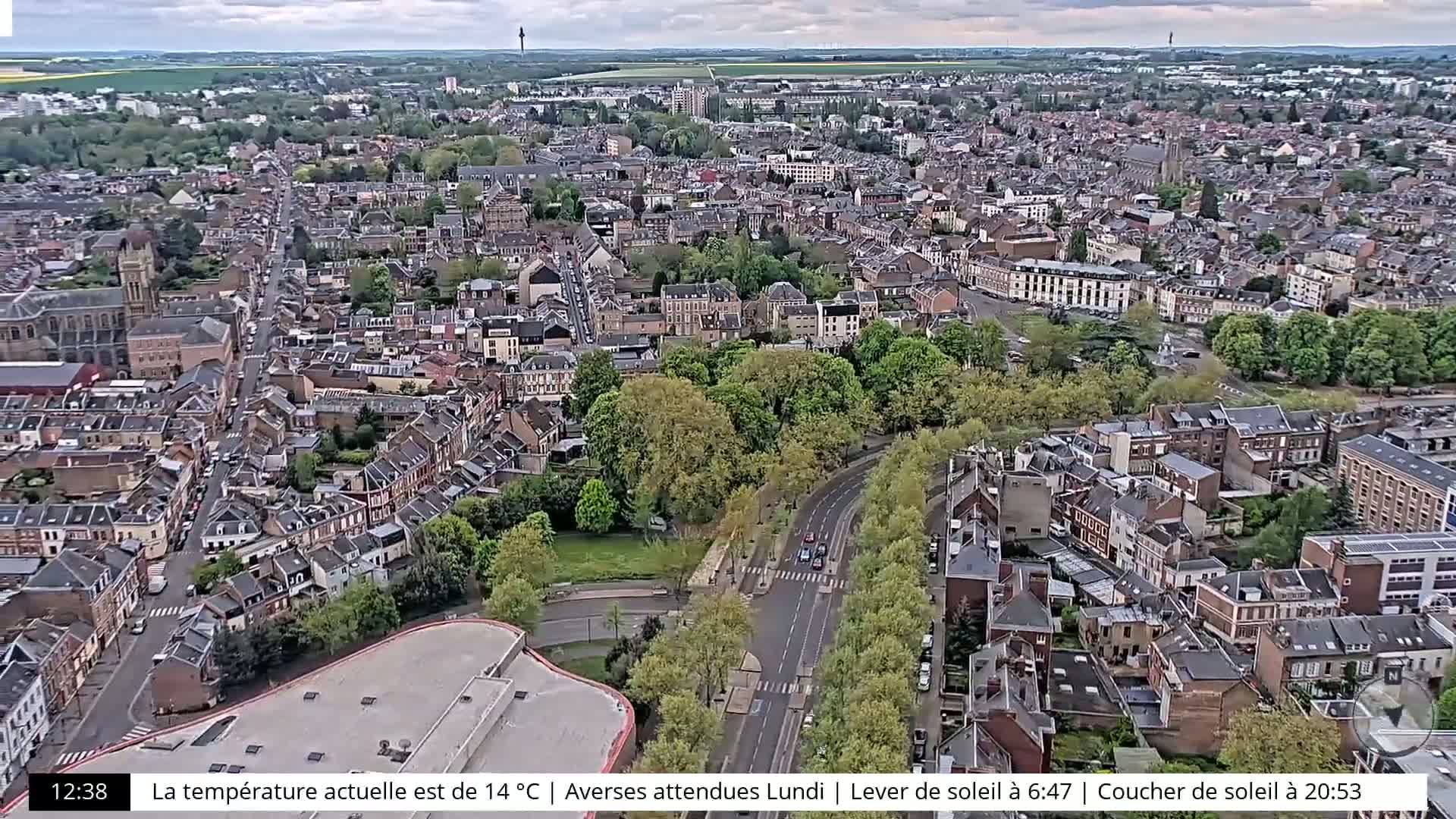 An aerial view of a city on an overcast day shows a large road cutting through a densely populated area with many trees and buildings.