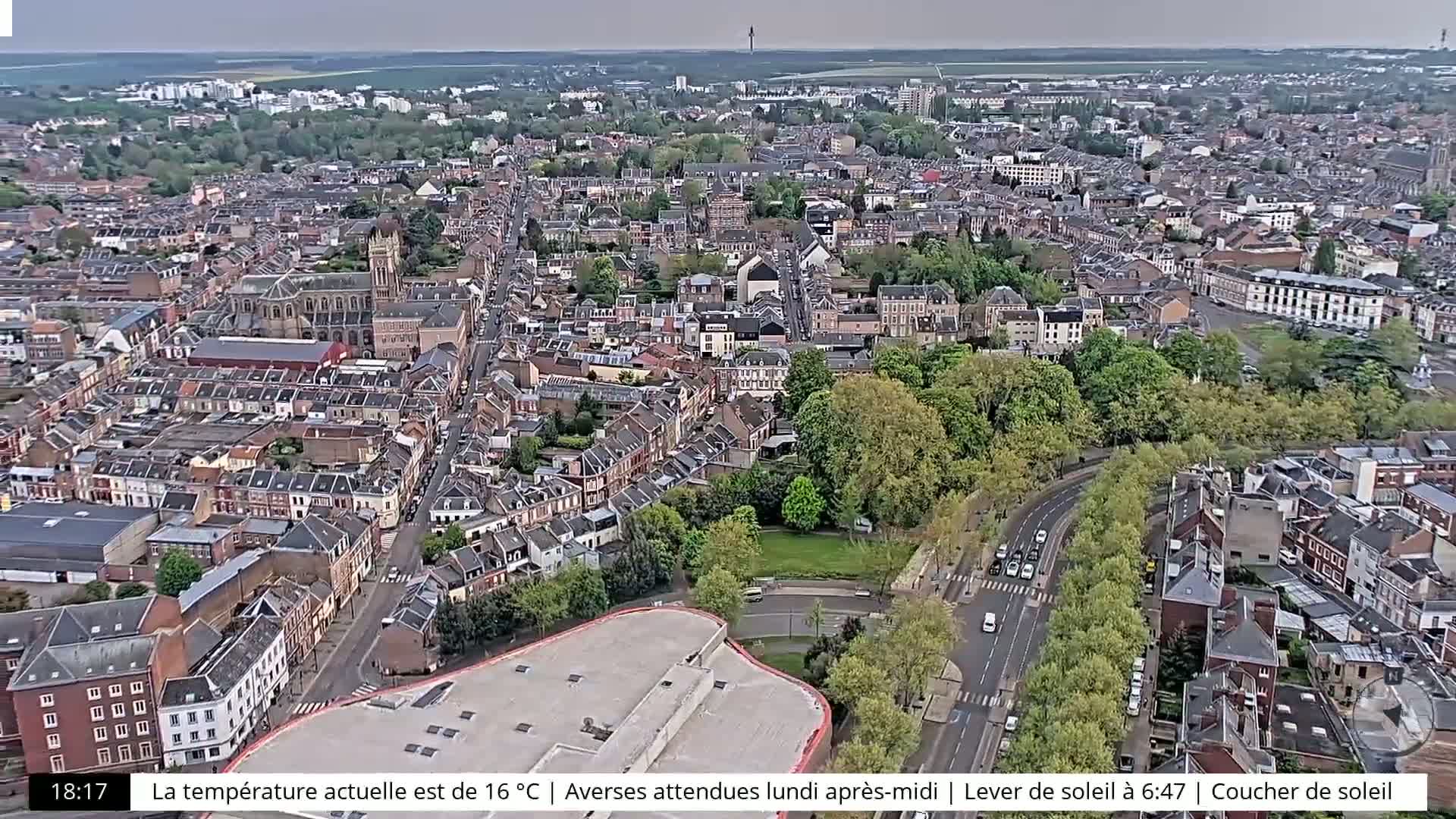 A high-angle, long shot of a city on an overcast day shows many buildings, roads, and green spaces.