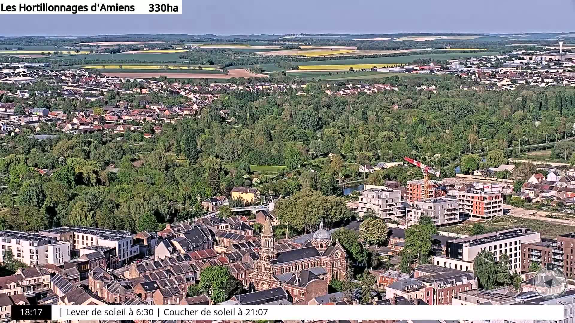 A high-angle, long shot on a sunny day shows a town nestled among lush green trees and fields, with a partially constructed building complex visible in the foreground.