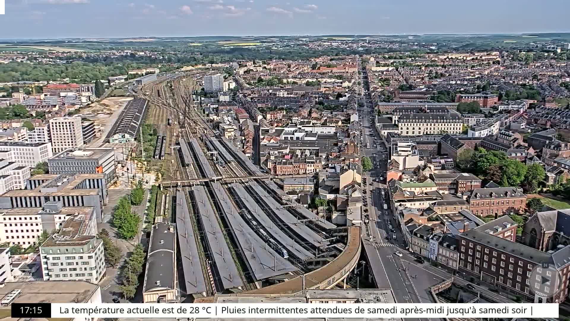 An aerial view shows a city with a large train station, under partly cloudy skies.