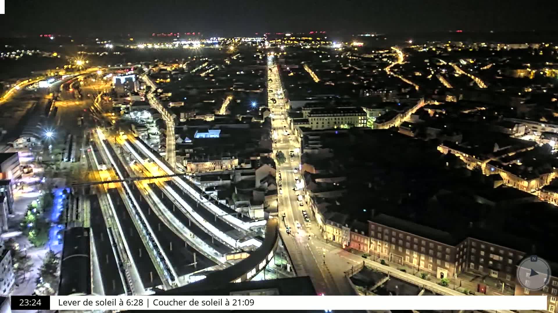A nighttime aerial view shows a city with illuminated streets and a train station, under a clear, dark sky.