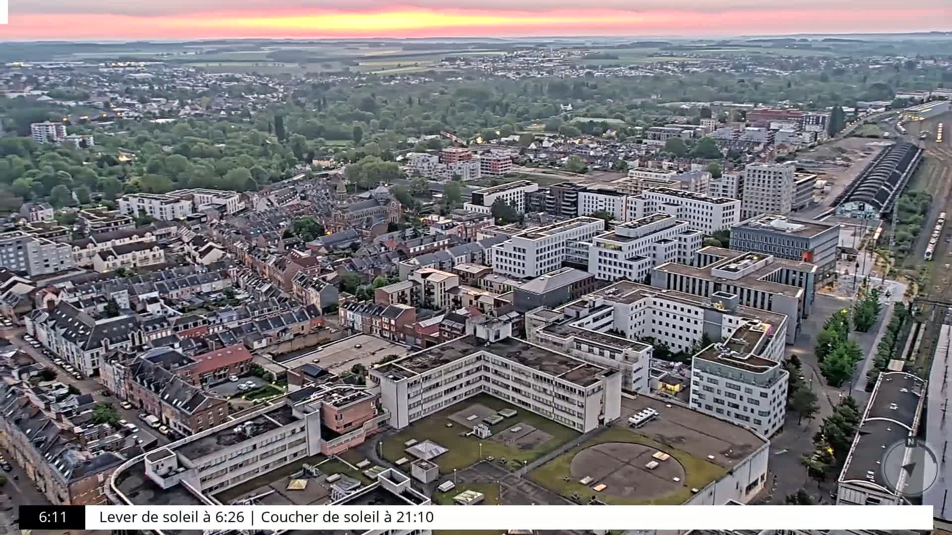 An aerial view shows a dense cityscape at sunrise, with many buildings and trees visible under a pinkish sky.