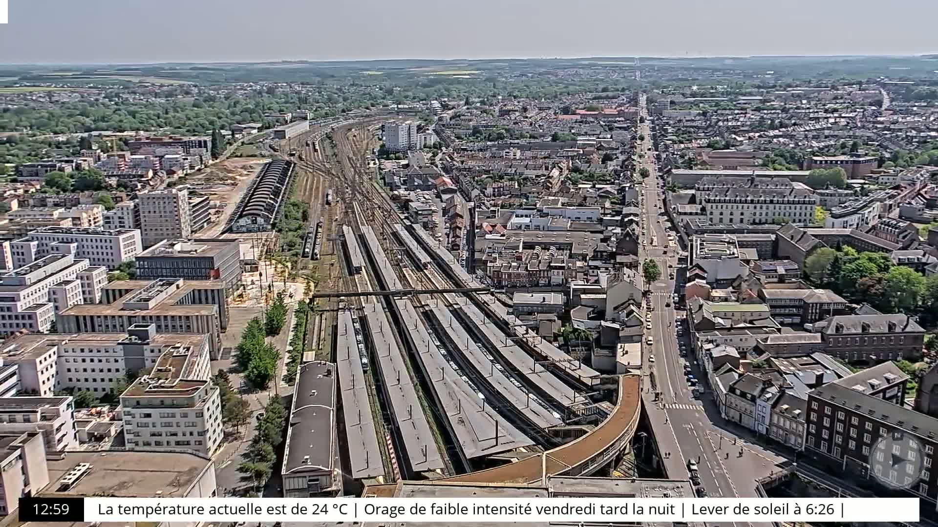 An aerial view of a city on a sunny day, showing a large train station with numerous tracks and platforms, surrounded by residential and commercial buildings.