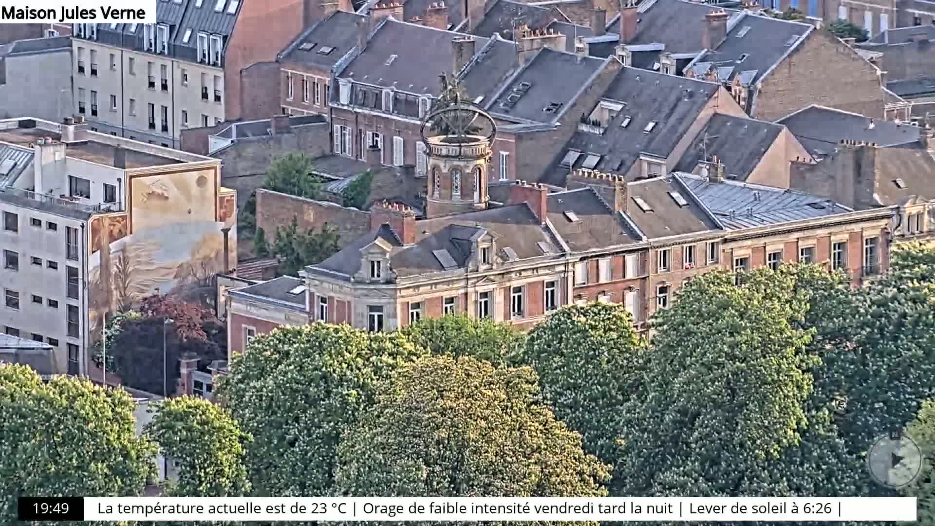 An aerial view shows a city block with red-brick buildings, a large mural on one building, and a tower with a weathervane, all partially obscured by green trees.