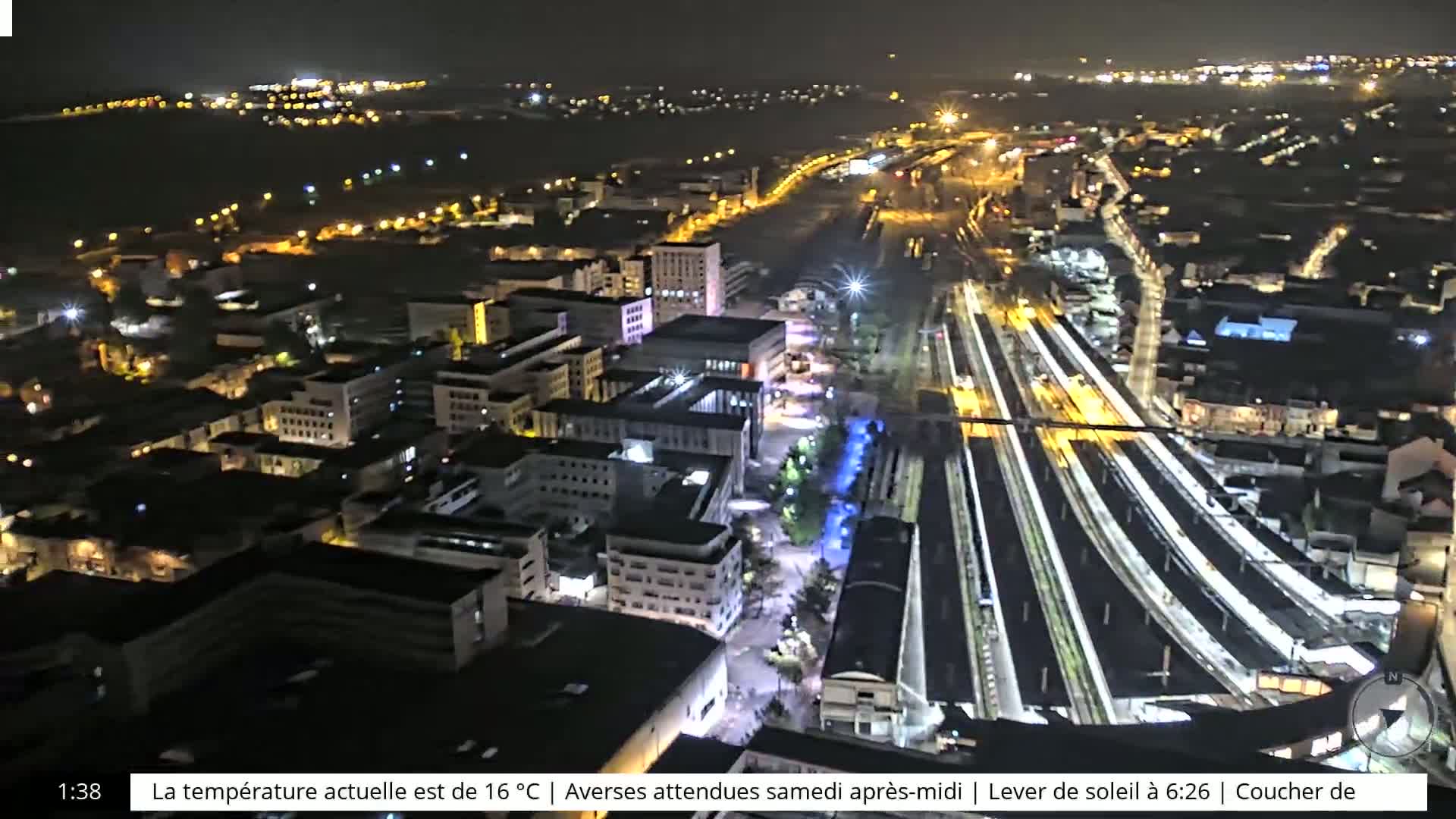 A nighttime aerial view shows a city with illuminated buildings and train tracks.