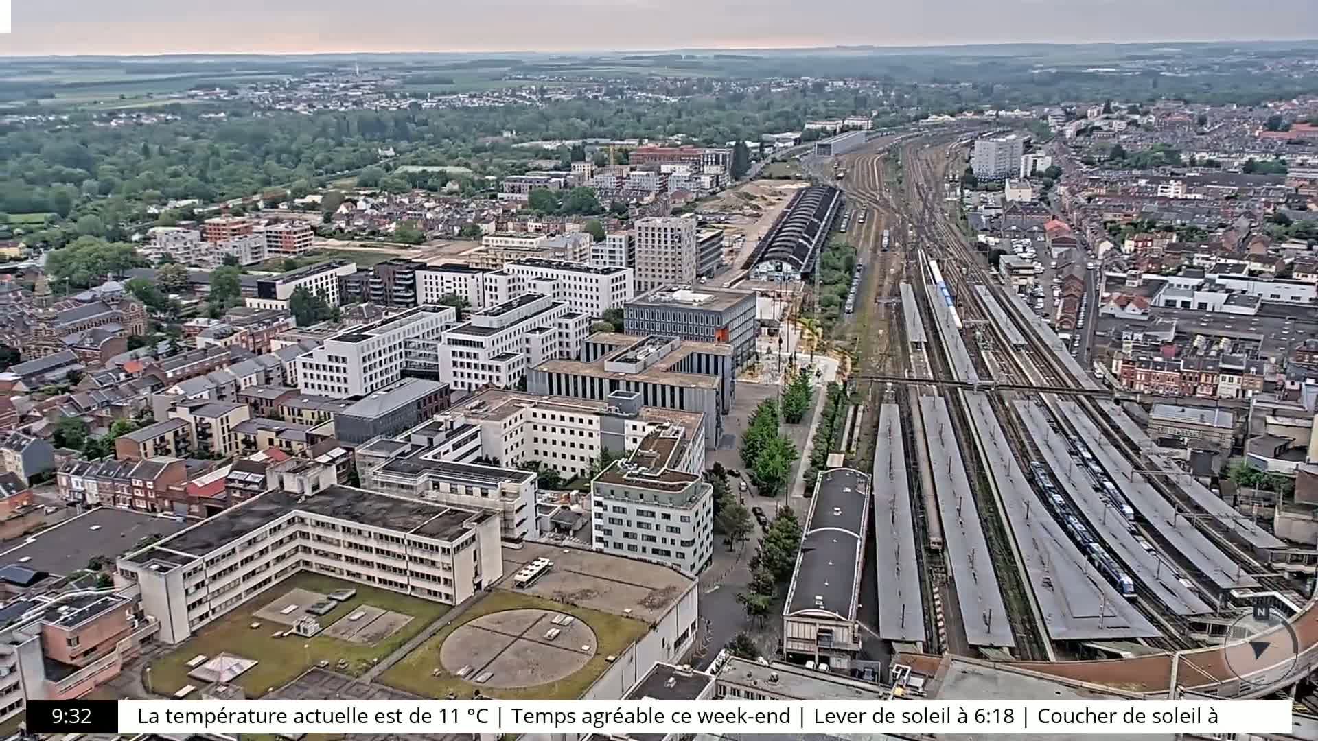 An aerial view shows a city with many buildings, a large train station, and surrounding green areas under an overcast sky.