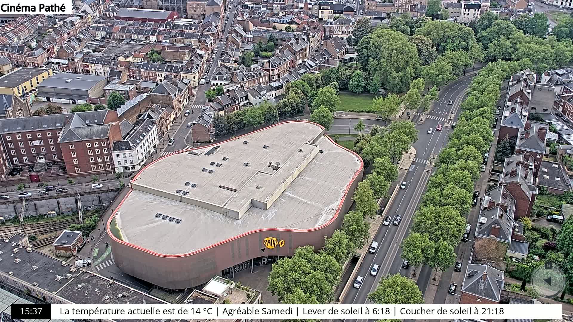 An aerial view shows a large, curved building surrounded by a city with tree-lined streets and sunny weather.