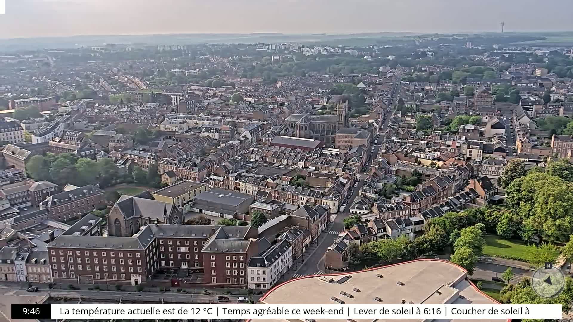 An aerial view of a city on a partly cloudy day, showing numerous buildings and green spaces.