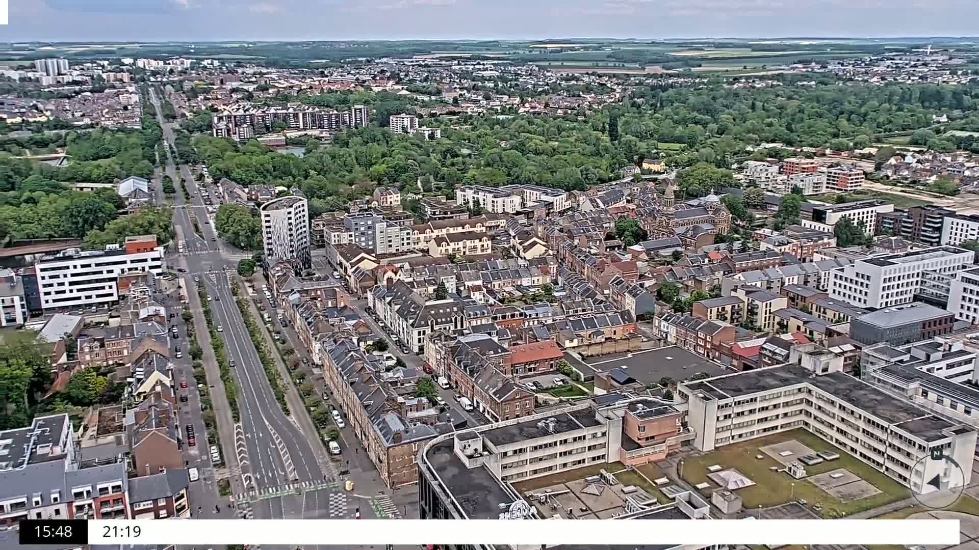 An aerial view shows a densely populated residential area with a long, straight road cutting through it under partly cloudy skies.