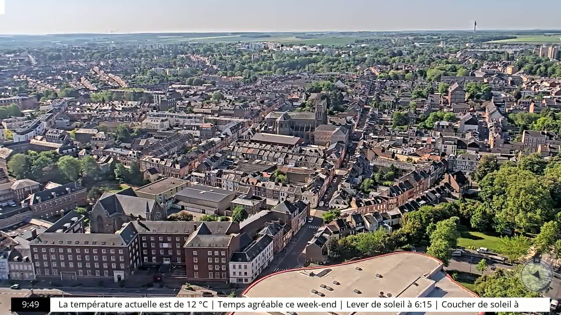 An aerial view shows a densely populated city on a sunny day, with many brick buildings and green spaces interspersed.