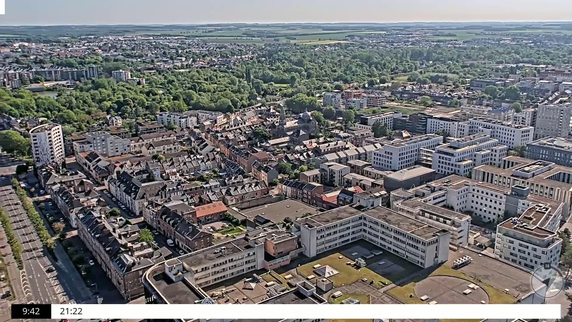 An aerial view shows a city under sunny skies, with a mix of residential buildings and larger structures, including a large complex of buildings that appear to be institutional in nature, surrounded by extensive green spaces.