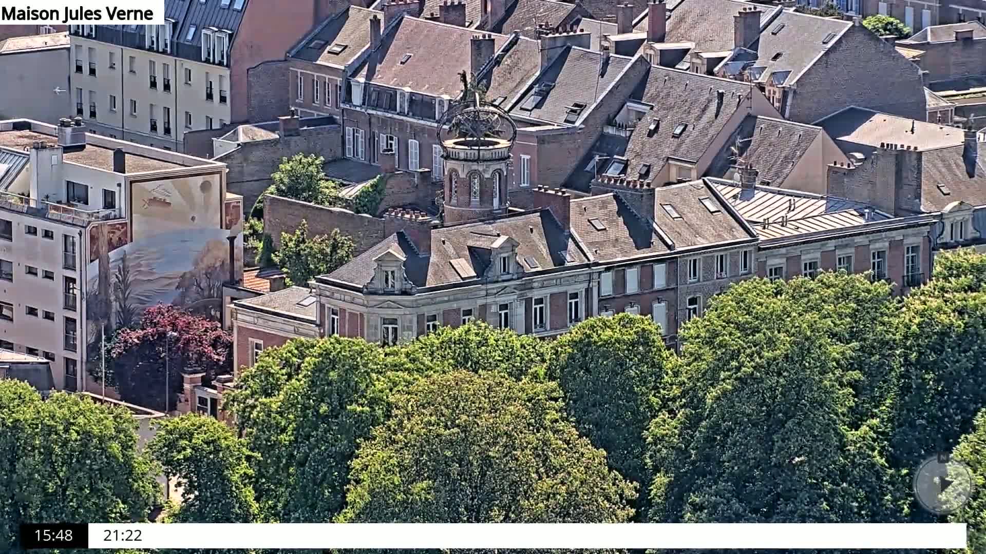 An aerial view shows a red-brick building with a unique tower, surrounded by lush green trees on a sunny day.