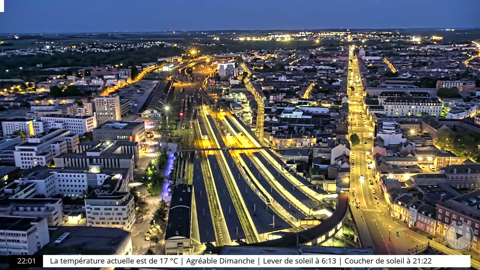 An aerial nighttime view of a city illuminated by streetlights, featuring a brightly lit train station at the center.