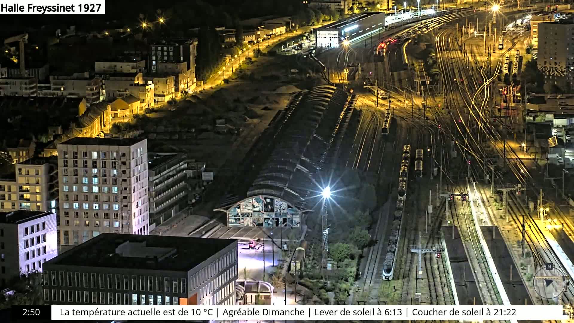 A nighttime aerial view of a train station and surrounding city, with many buildings illuminated and trains visible on numerous tracks.
