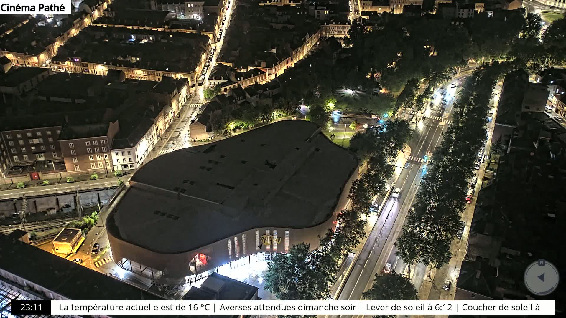 An aerial nighttime view of a city shows a large, curved building next to a wet street lined with trees and cars.