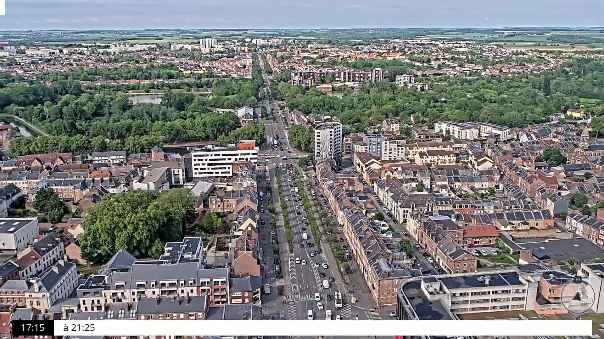 An aerial view shows a city on a partly cloudy day, with a main road lined with trees running through a densely populated area of primarily brick buildings.