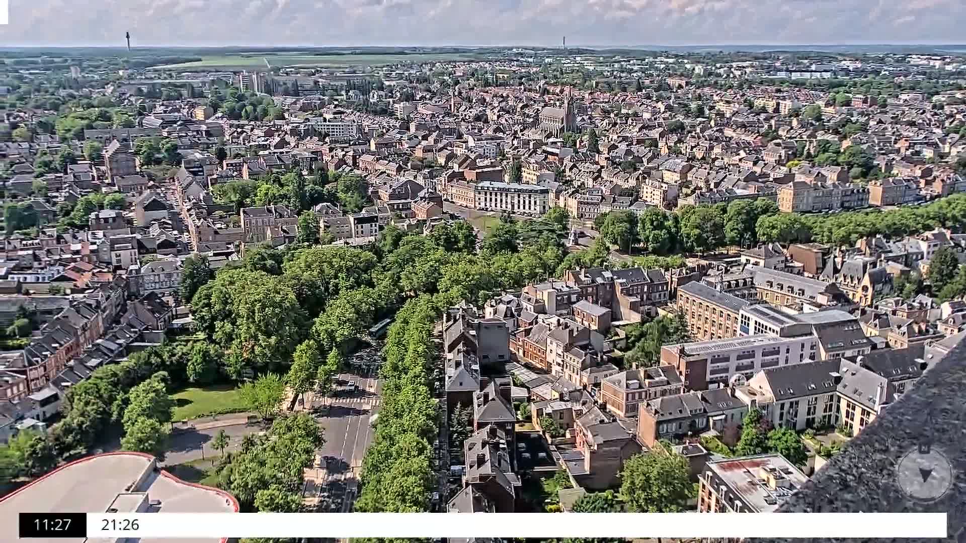 An aerial view shows a sprawling city on a partly cloudy day, with numerous buildings nestled among green trees and parks.