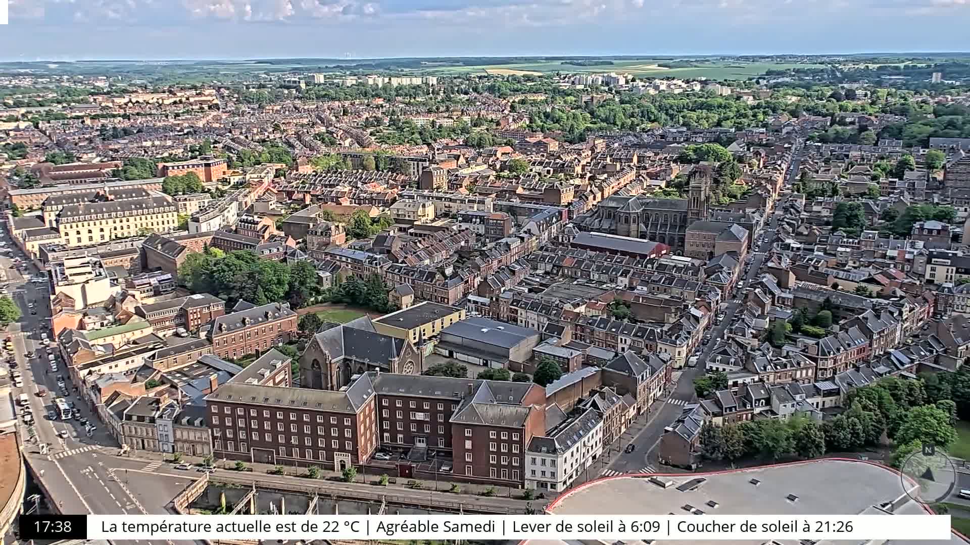 An aerial view of a densely packed European city on a partly sunny day, showing many brick buildings and tree-lined streets.