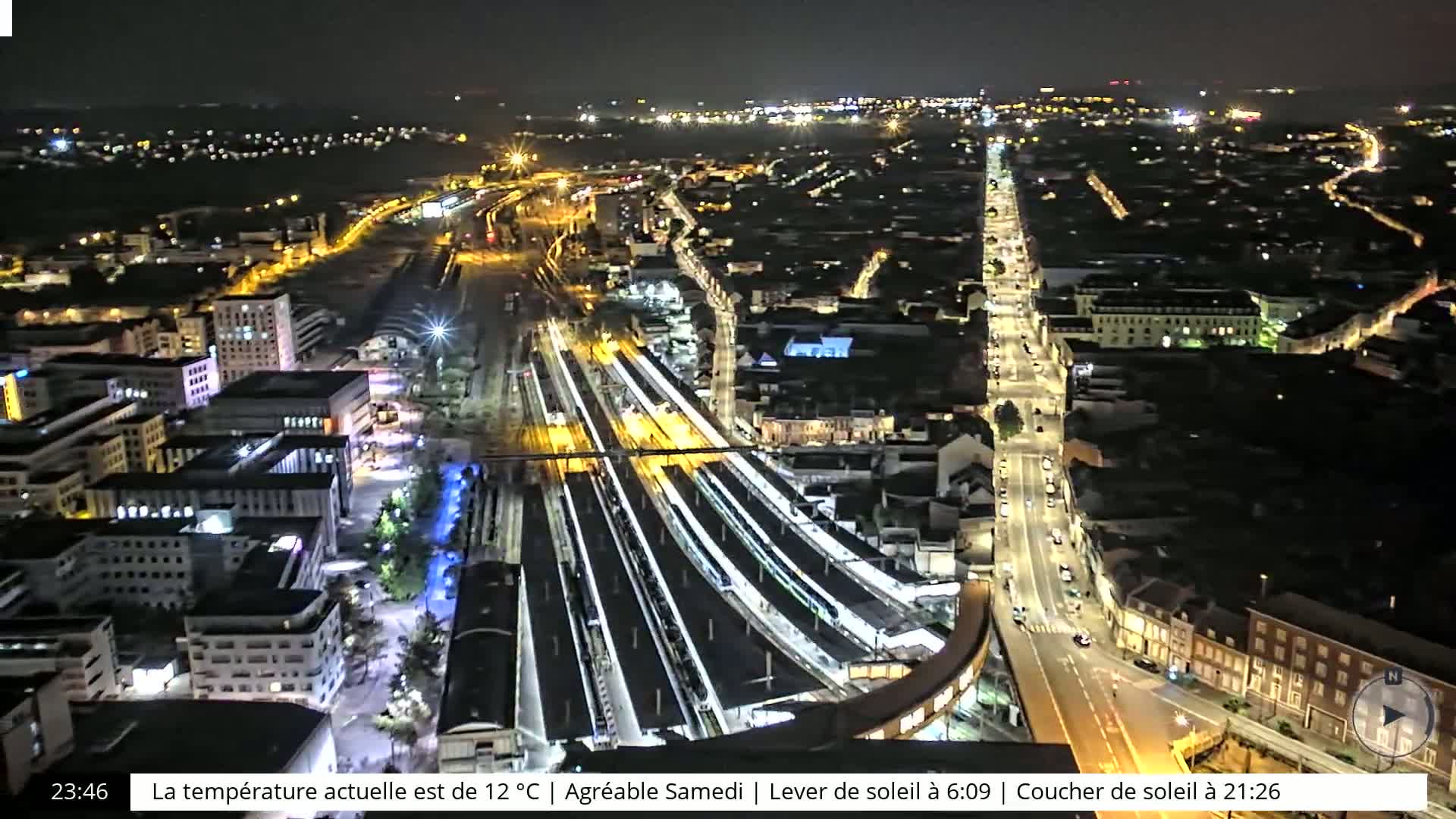 A nighttime aerial view of a city with a brightly lit train station and road, under a clear sky.