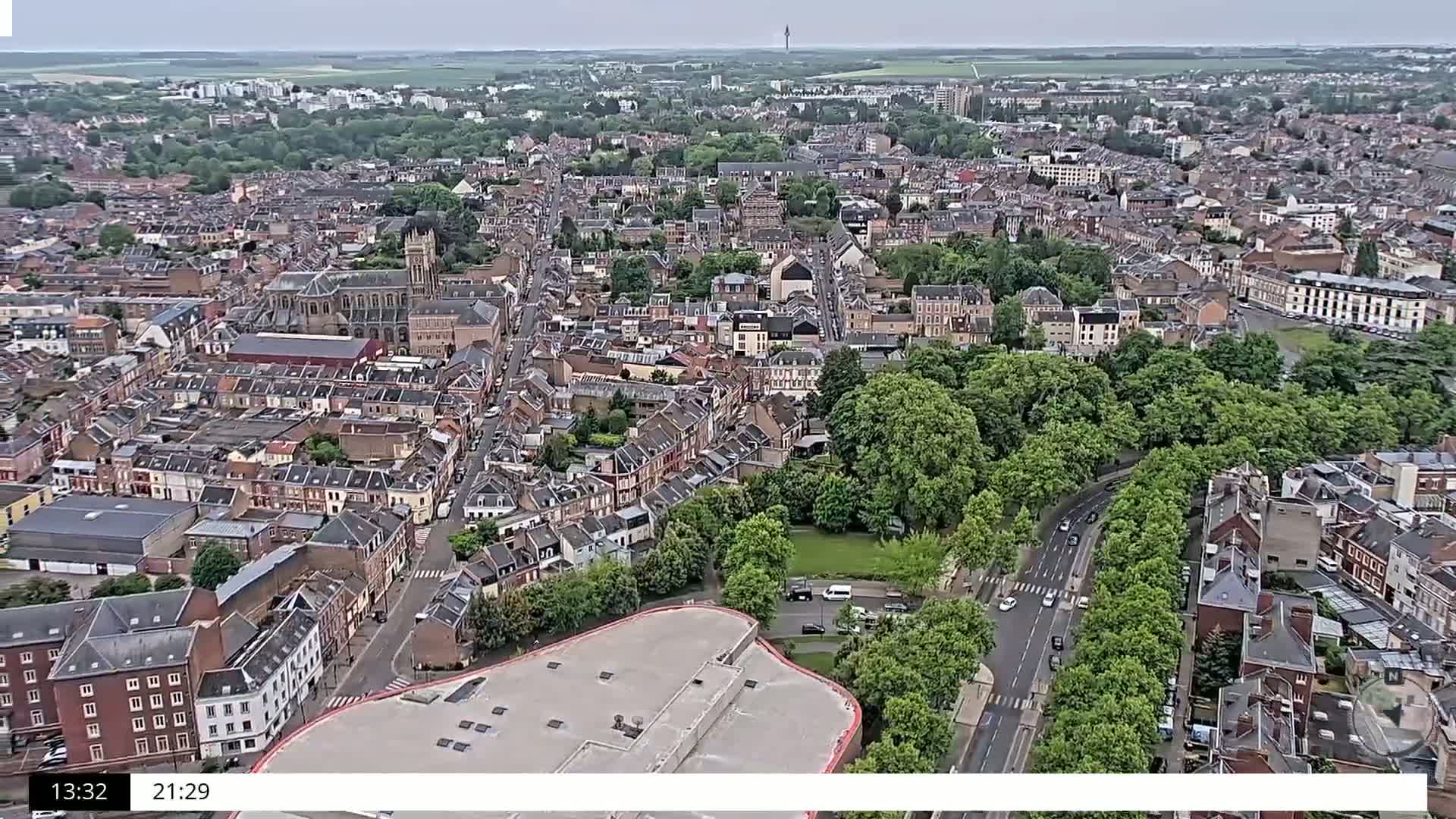 An aerial view shows a densely packed town on an overcast day, with a large park and a road cutting through the green space.