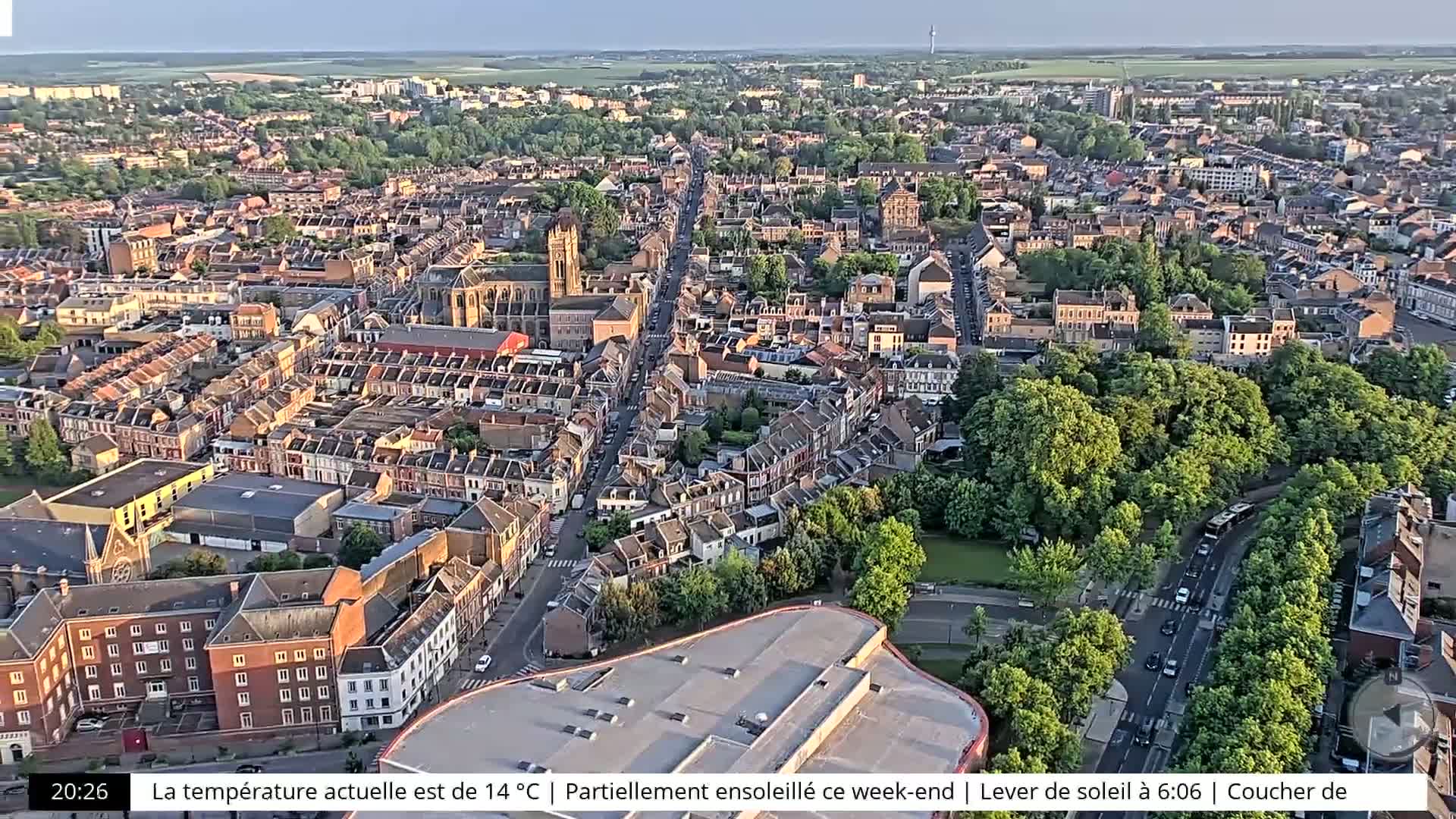 An aerial view of a town shows many closely packed buildings surrounded by lush green trees under partly sunny skies.