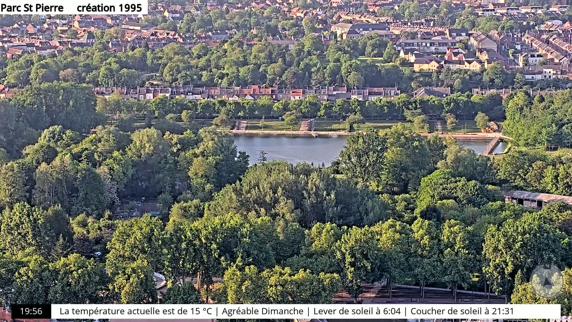 An aerial view shows a park with a pond surrounded by lush green trees and residential buildings in the background, under sunny conditions.