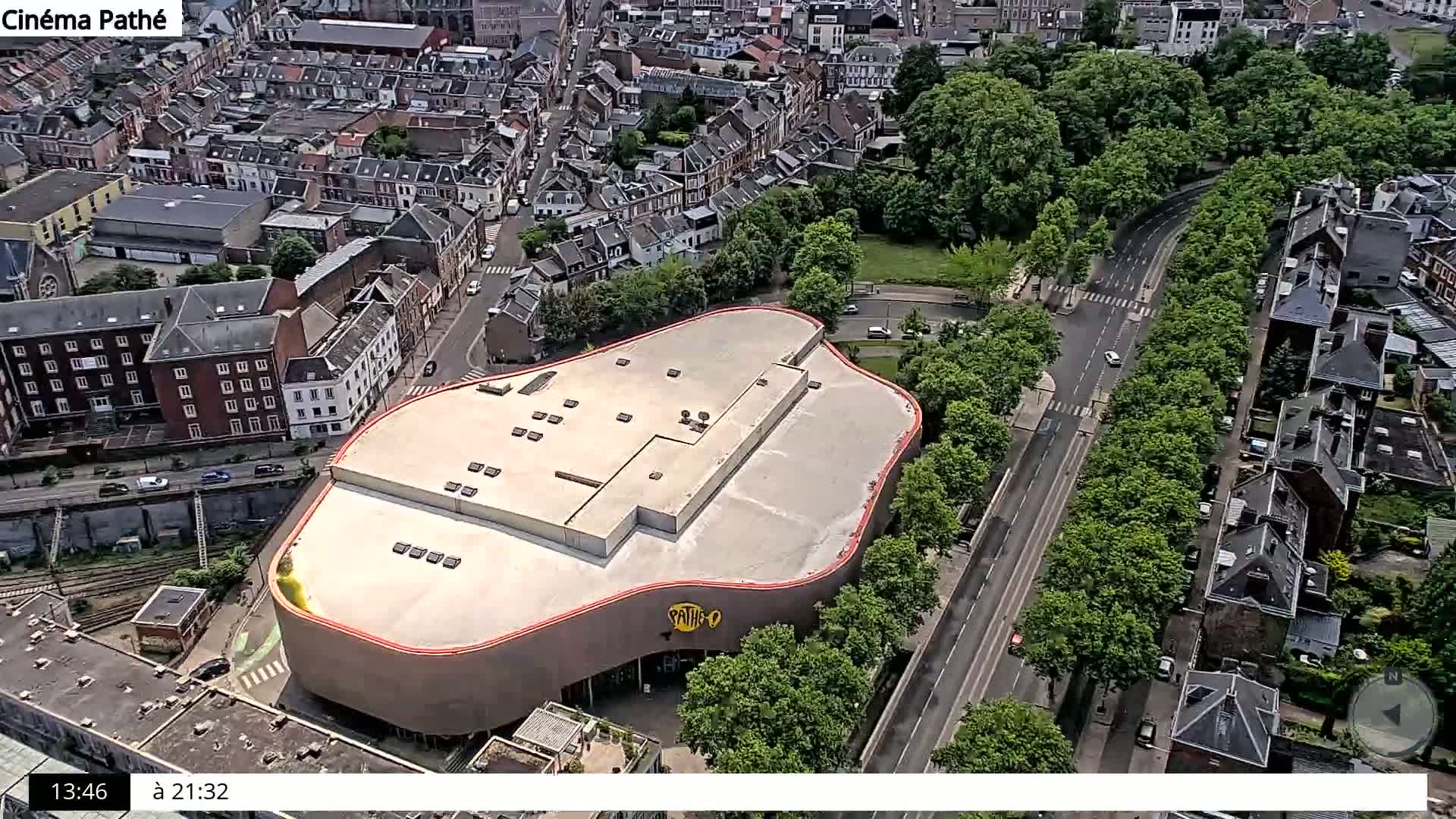 An aerial view shows a city block with a large, uniquely shaped building, surrounded by residential buildings, a tree-lined road, and a park on a sunny day.