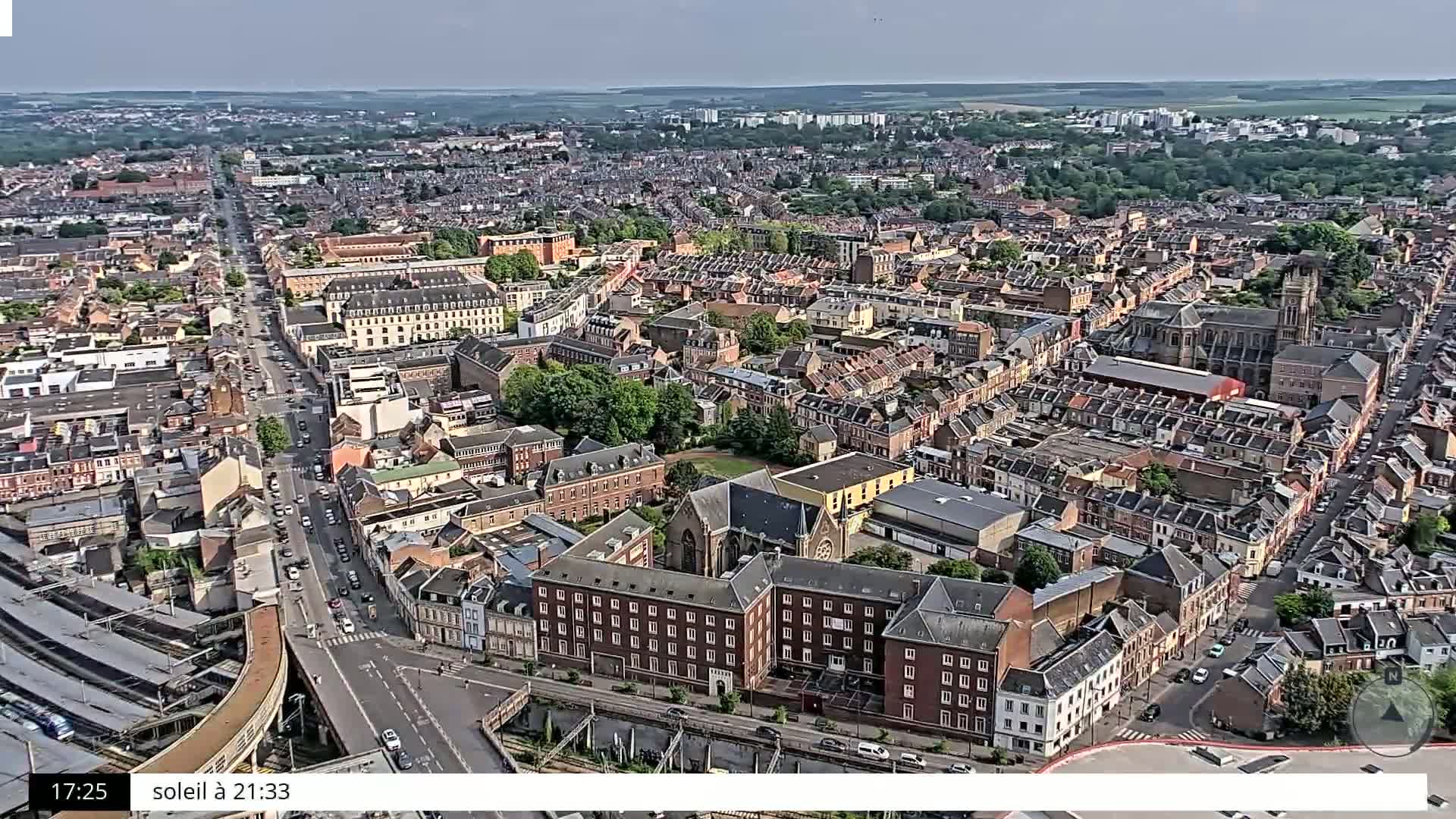 A high-angle, sunny view of a densely populated European city with a mix of residential and commercial buildings, a church, and a railway line.