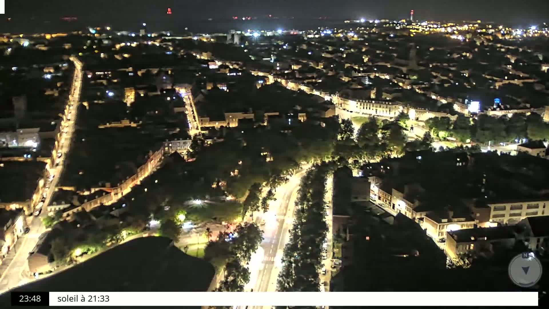 A nighttime aerial view of a city with illuminated streets and buildings, surrounded by dark green spaces.
