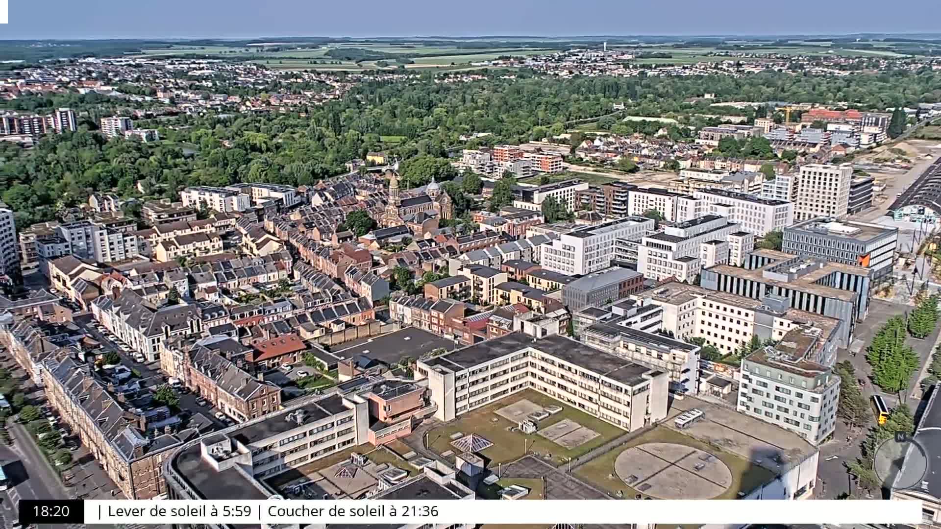 An aerial view shows a dense collection of buildings in a city on a clear, sunny day, surrounded by green trees and open spaces.