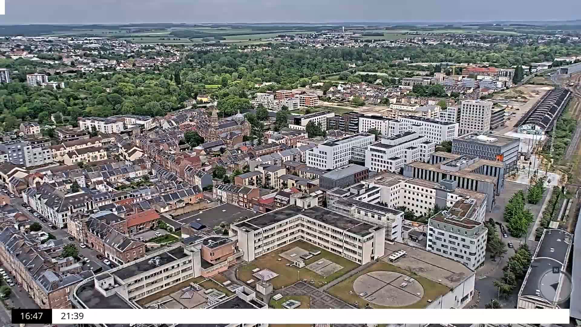 An aerial view of a city on an overcast day shows a mix of residential buildings and modern structures, with a large complex in the foreground and surrounding green spaces.