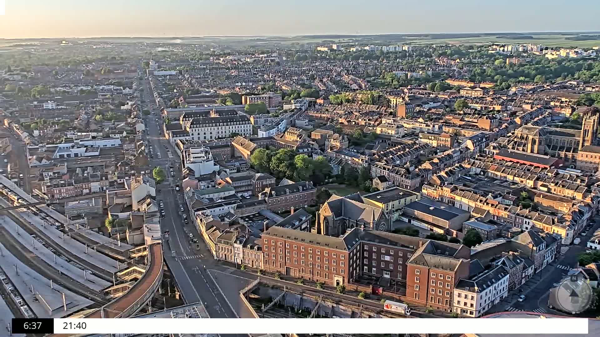 An aerial view of a densely populated city, under a clear, bright sky at sunrise, shows a long, straight road cutting through the city, alongside train tracks and a highway.