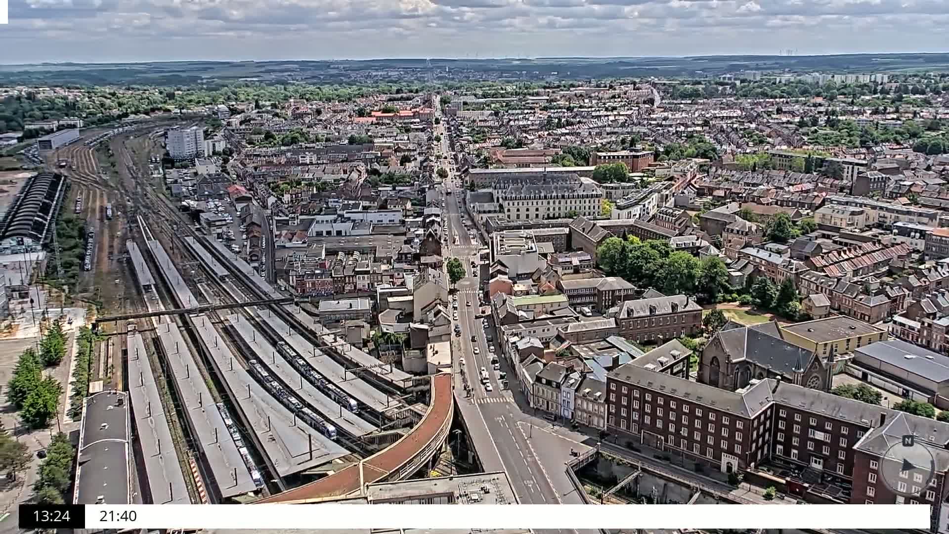 A high-angle, long shot shows a sprawling city under partly cloudy skies, with a large train station and many parallel train tracks visible in the foreground.