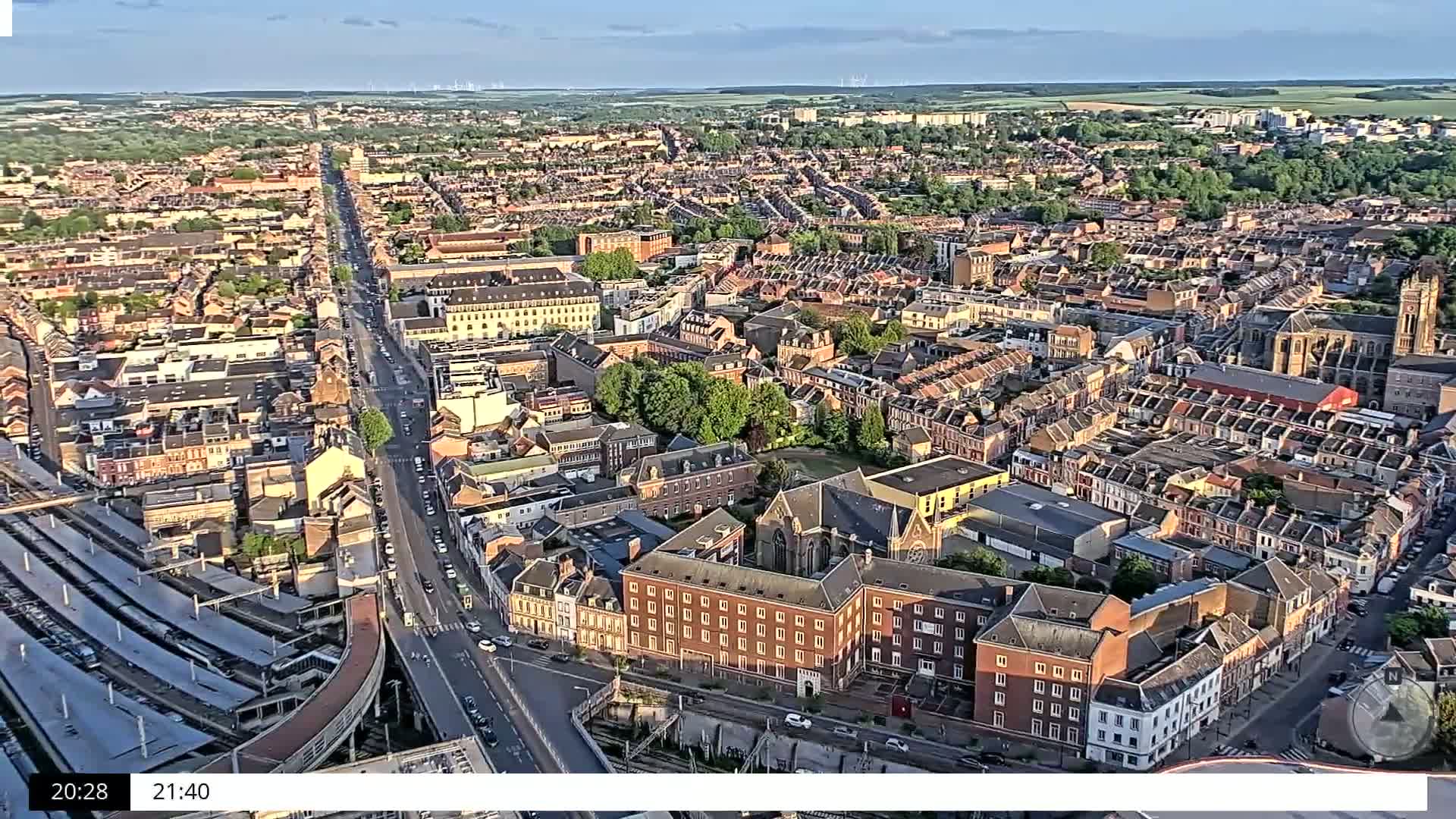 An aerial view shows a densely populated city under a partly cloudy, late afternoon sky, with a long, straight road running through the center.