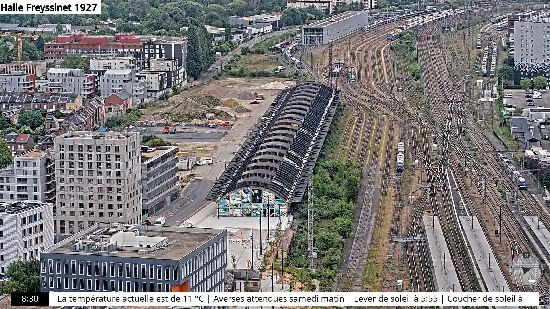 An aerial view shows a large train yard with numerous tracks and parked trains, adjacent to a city with modern and older buildings under partly cloudy skies.