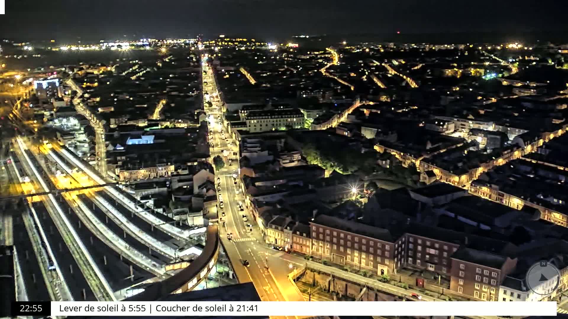 An aerial nighttime view of a city with illuminated streets and buildings, featuring train tracks in the foreground.