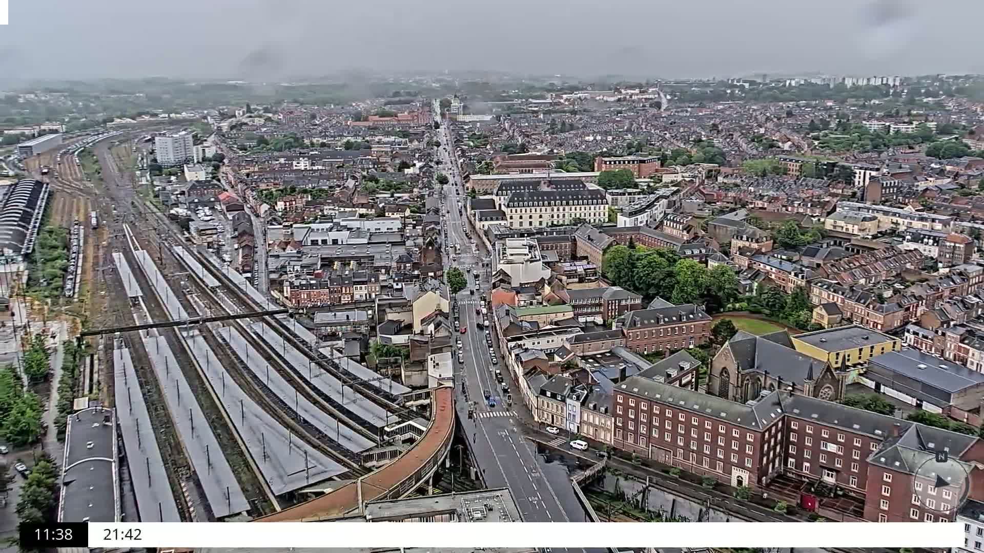An aerial view of a city on an overcast day shows a train station, a long road, and many buildings of varying sizes and styles.