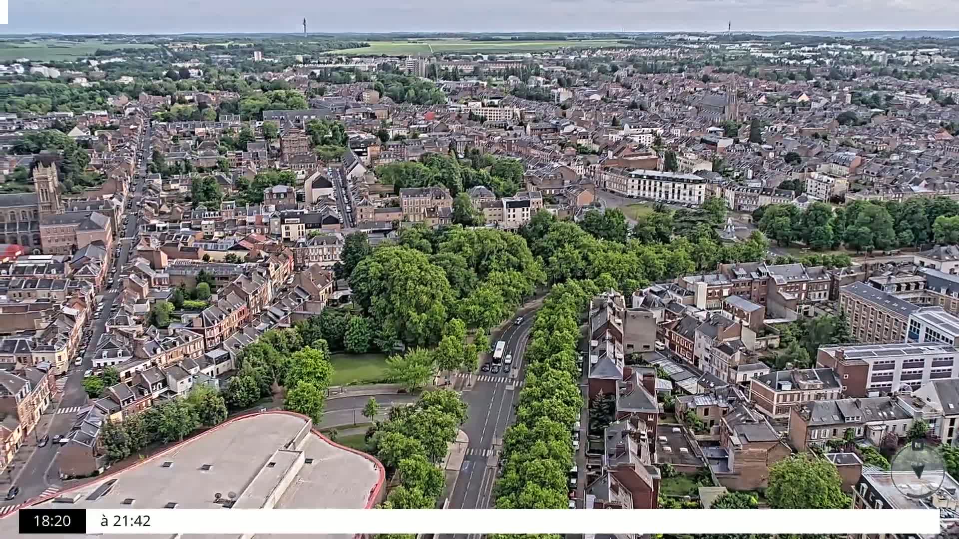 An aerial view shows a city under partly cloudy skies, with a large park nestled among numerous closely-packed buildings.