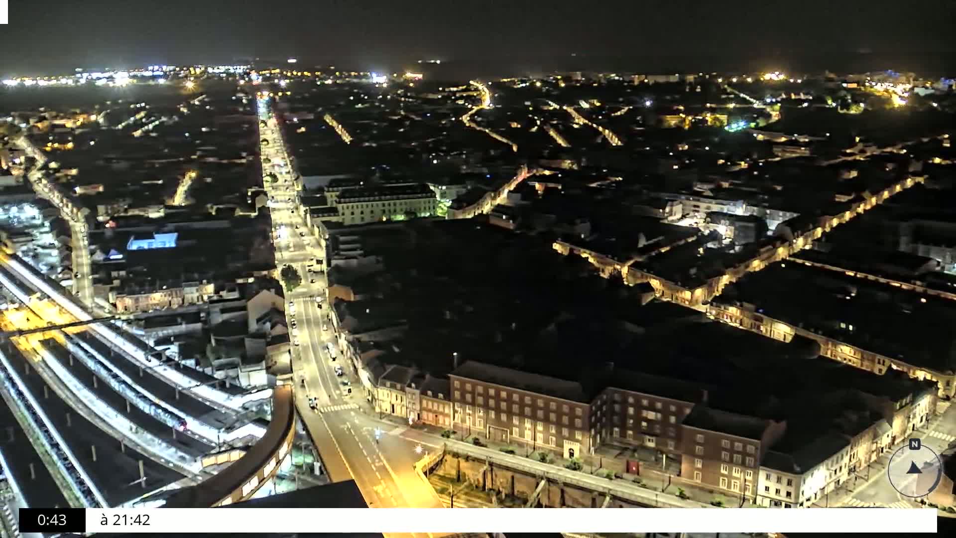 A nighttime aerial view of a city with illuminated streets and buildings, showing a train station and surrounding residential areas.