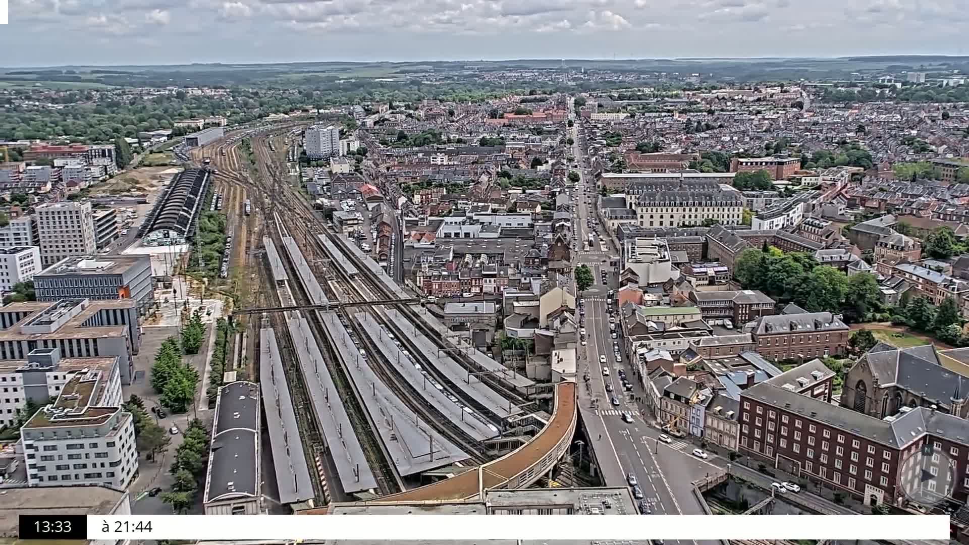 A sprawling cityscape under a partly cloudy sky features a large train station with numerous tracks and platforms, surrounded by residential and commercial buildings.