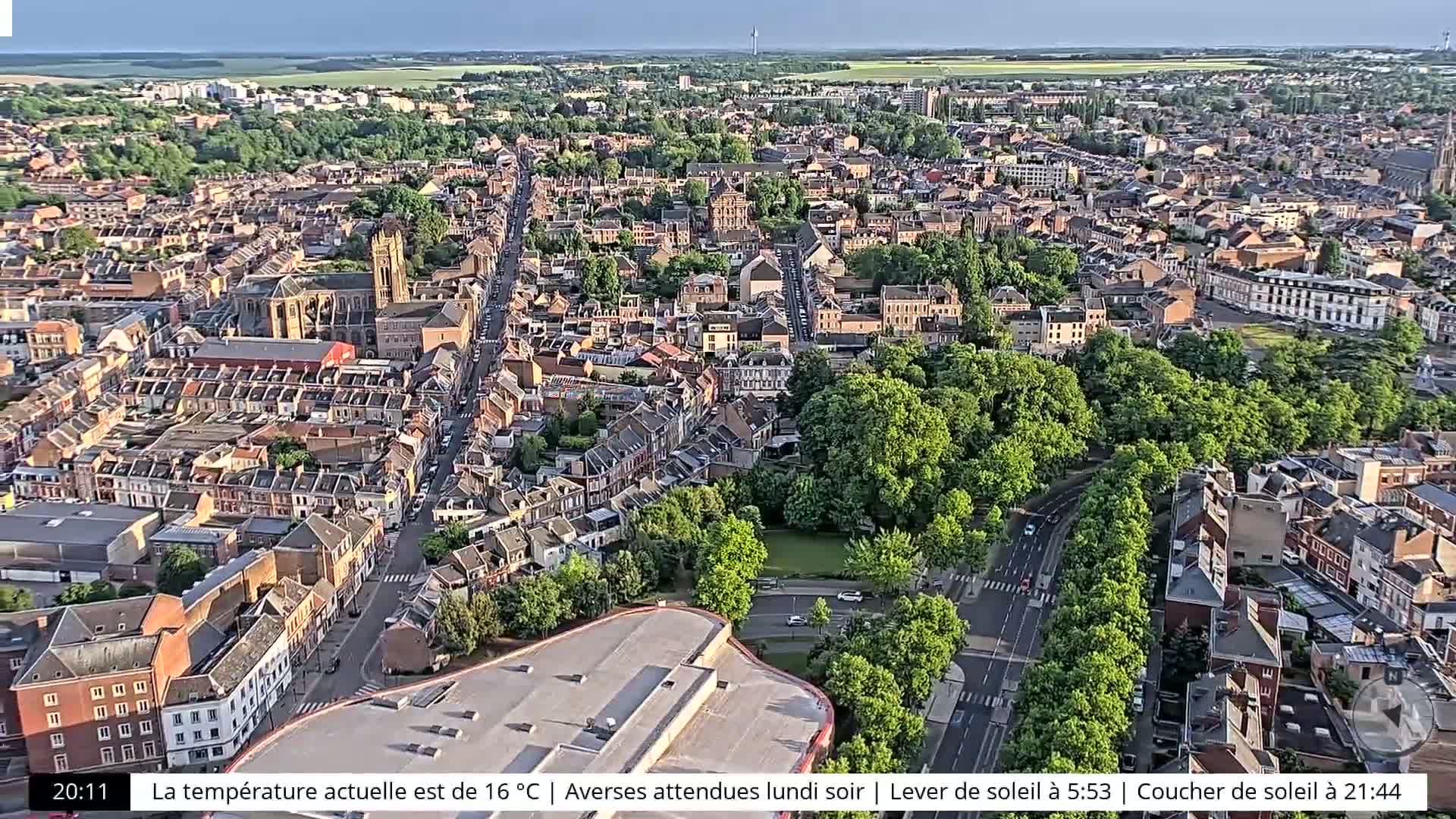 An aerial view of a city with numerous buildings, tree-lined streets, and a large park under a partly cloudy sky.