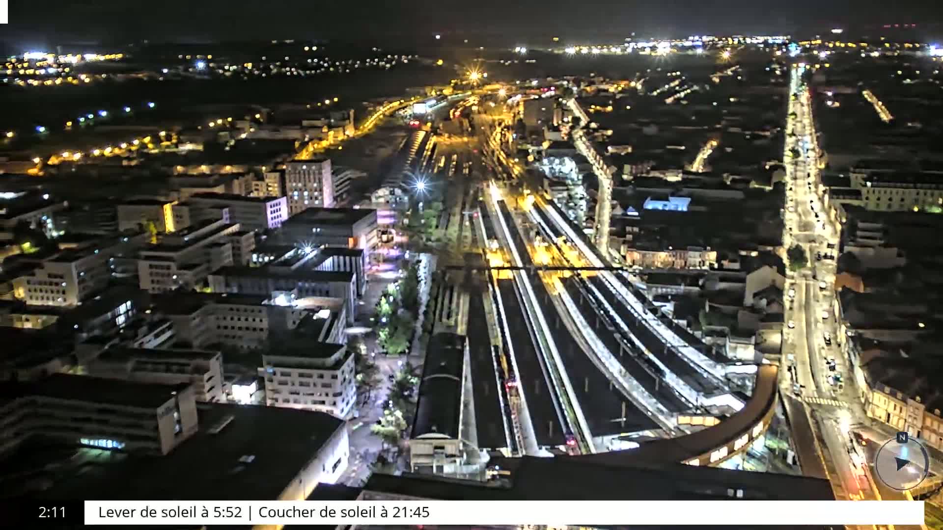 A nighttime aerial view of a city showing a brightly lit train station and surrounding buildings and streets.