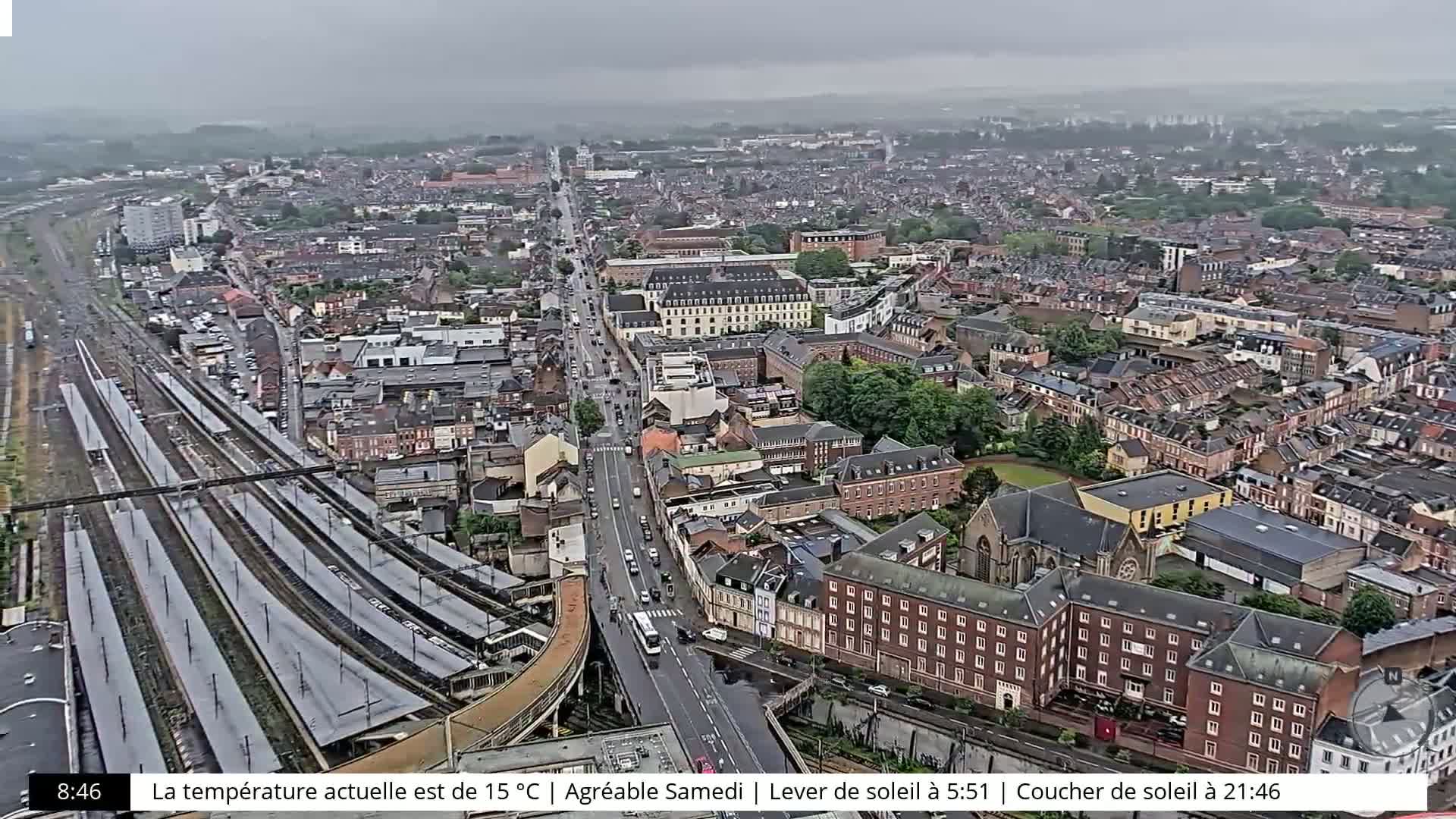 A high-angle, long shot of a city on an overcast day shows a train station, roads, and many buildings.
