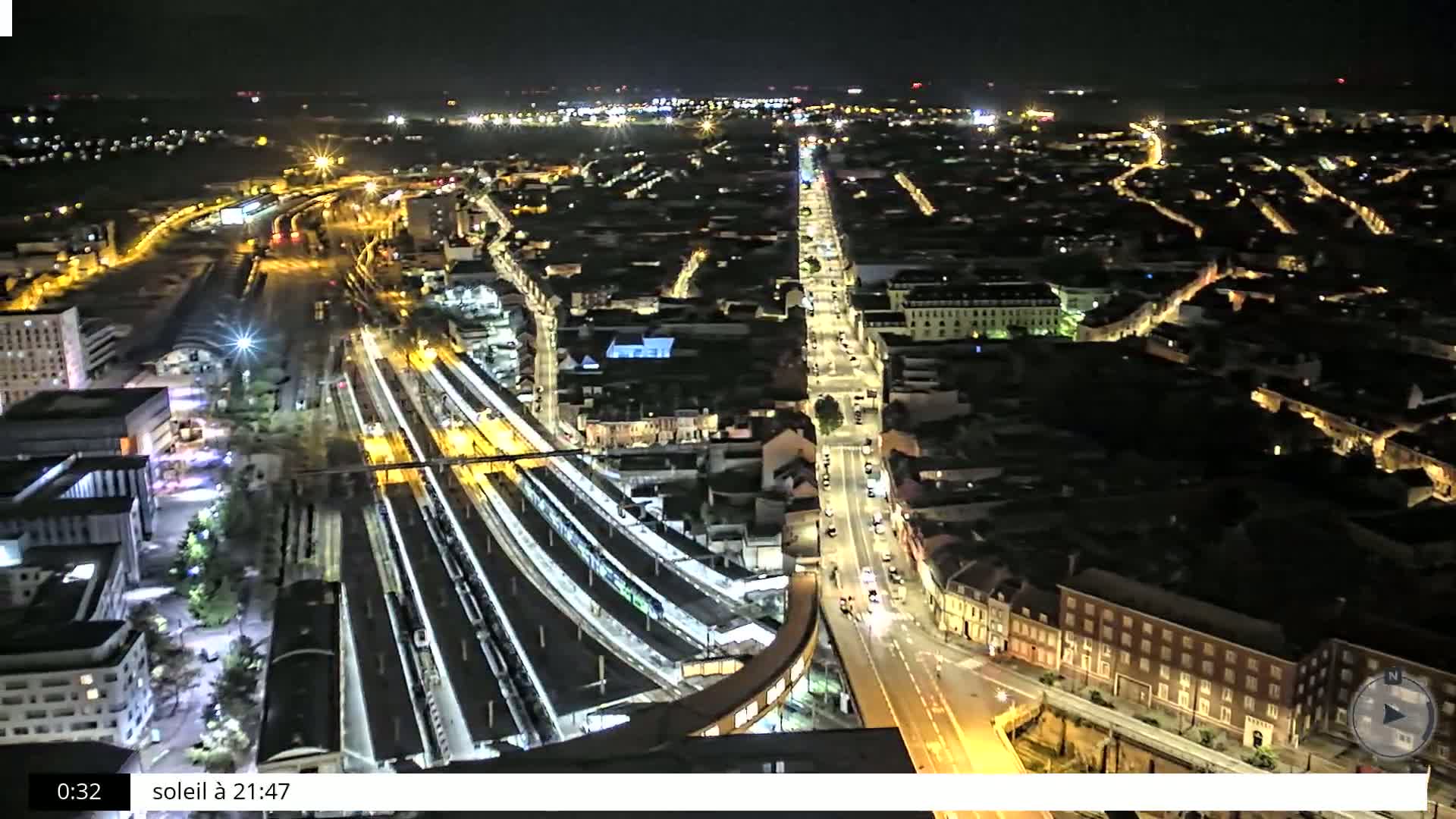 A nighttime aerial view shows a city with a train station and illuminated streets.
