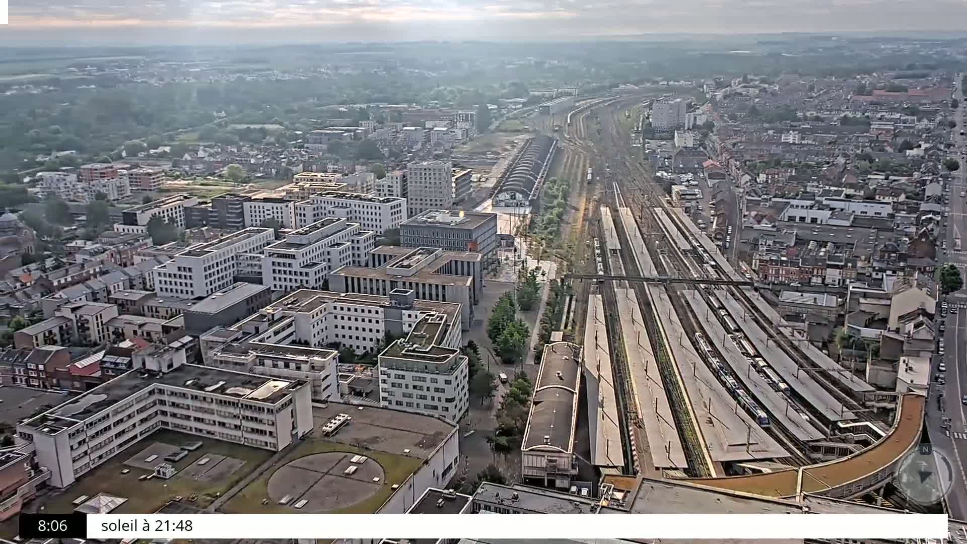 An aerial view of a city on a partly sunny day shows a large train station with many tracks and trains, surrounded by numerous buildings of varying heights.