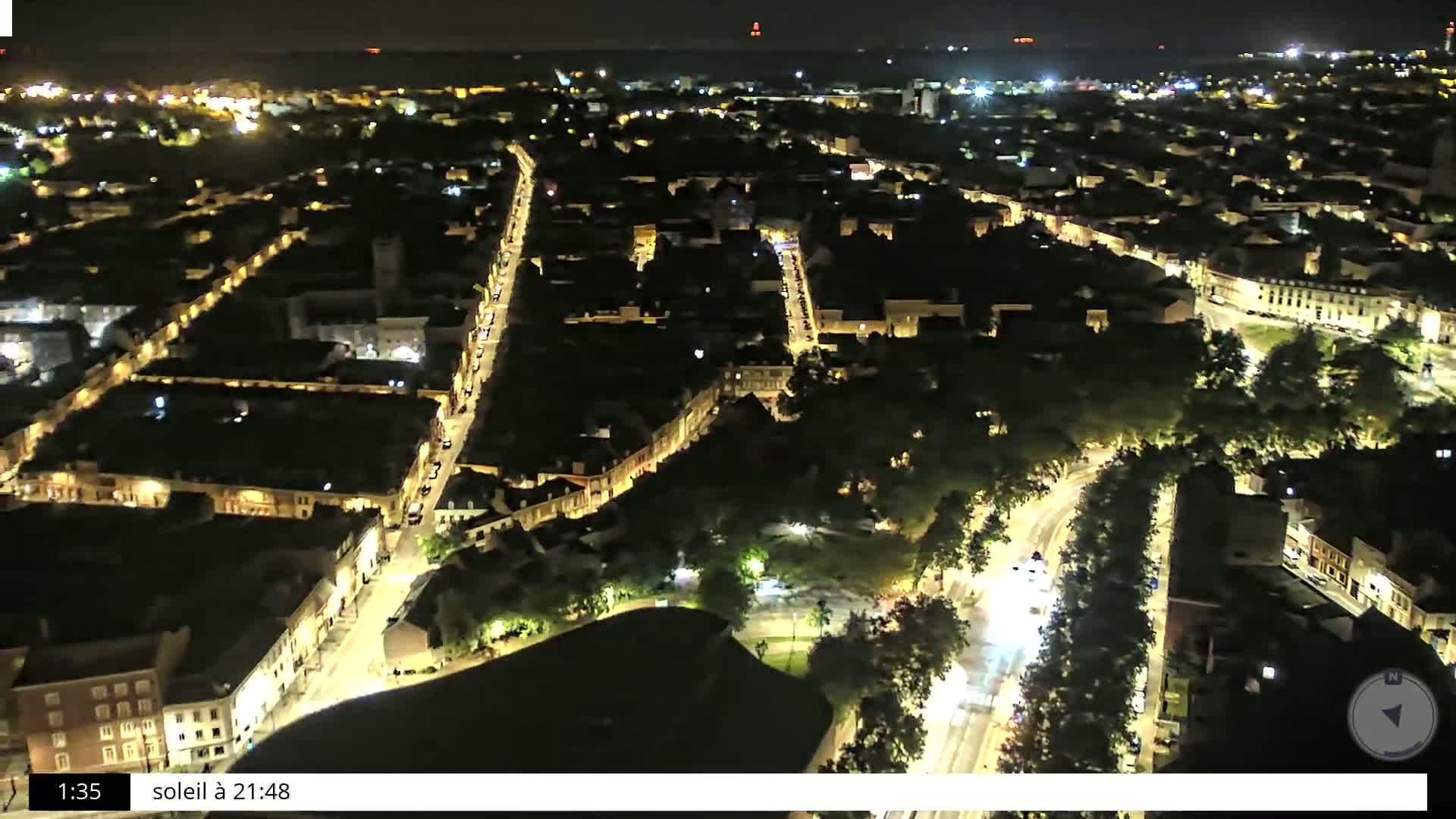 A nighttime aerial view shows a city with illuminated streets and buildings surrounded by dark green trees.